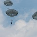The image shows multiple military paratroopers descending from the sky using round, olive-drab/gray military parachutes. At least five parachutes are visible at varying altitudes against a partly cloudy blue sky. The parachutes appear to be standard military static-line round canopies, consistent with airborne operations.