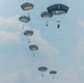 A military mass parachute jump is captured mid-air, showing approximately 10-12 paratroopers descending with round military-style parachutes against an overcast sky. The parachutes are gray/olive-drab in color, consistent with military static-line deployment canopies. The jumpers appear to be in a staggered vertical formation, suggesting they exited an aircraft in rapid succession.