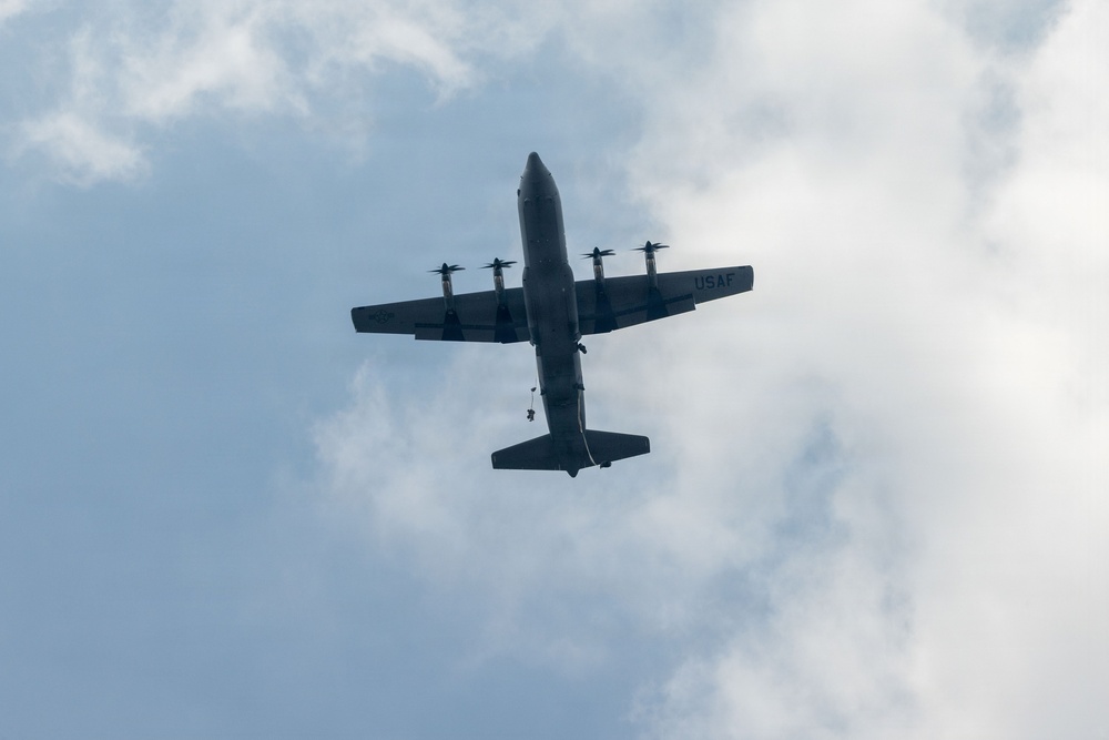 A USAF (United States Air Force) military transport aircraft, identifiable as a C-130 Hercules, is photographed from below against a partly cloudy sky. The aircraft appears to have a figure or object dangling beneath its fuselage, suggesting a parachute drop or airborne operation in progress. The image is taken from a ground-level upward perspective, showing the underside of the aircraft.