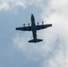 A USAF (United States Air Force) military transport aircraft, identifiable as a C-130 Hercules, is photographed from below against a partly cloudy sky. The aircraft appears to have a figure or object dangling beneath its fuselage, suggesting a parachute drop or airborne operation in progress. The image is taken from a ground-level upward perspective, showing the underside of the aircraft.