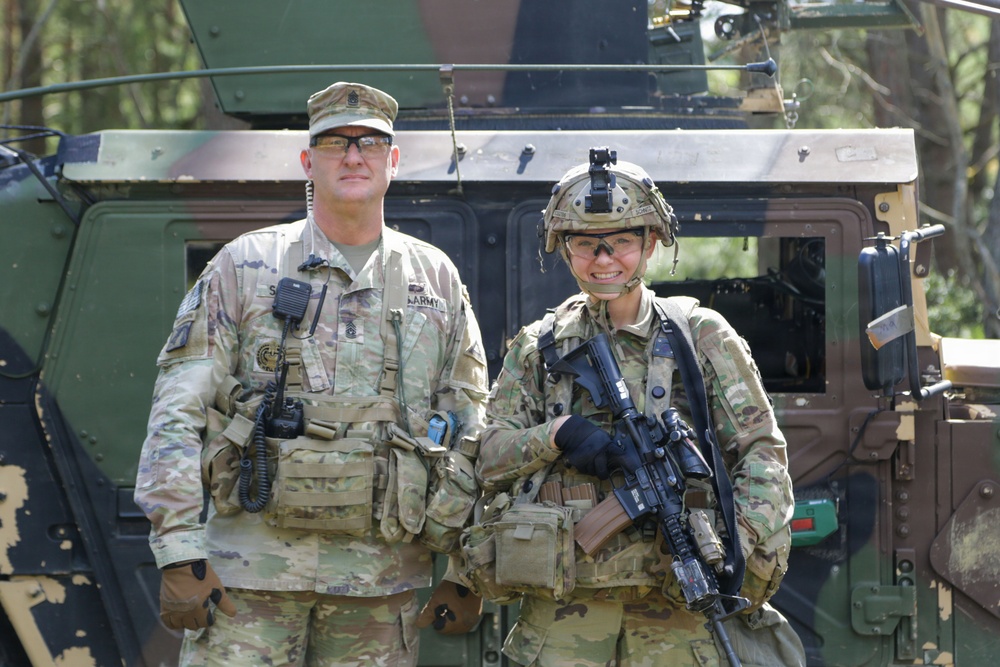 Two U.S. Army soldiers in MultiCam (OCP) camouflage uniforms are posing in front of a military vehicle, likely a Humvee or similar tactical vehicle. The soldier on the left wears a patrol cap and appears to be a senior NCO based on visible rank insignia, while the soldier on the right wears a combat helmet with night vision mount and carries an assault rifle. Both are smiling for the photo in what appears to be a field training or operational environment.