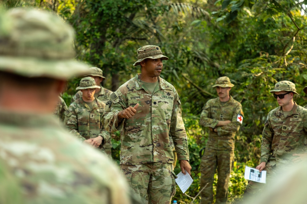 A U.S. Army officer identified by name tape as 'STEPPE' addresses a group of soldiers in an outdoor, jungle-like setting. He appears to be conducting a briefing or after-action review, holding papers and gesturing while speaking. Several other soldiers in OCP (Operational Camouflage Pattern) uniforms and boonie hats stand listening, including one with a visible Red Cross medical patch.