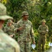 A U.S. Army officer identified by name tape as 'STEPPE' addresses a group of soldiers in an outdoor, jungle-like setting. He appears to be conducting a briefing or after-action review, holding papers and gesturing while speaking. Several other soldiers in OCP (Operational Camouflage Pattern) uniforms and boonie hats stand listening, including one with a visible Red Cross medical patch.