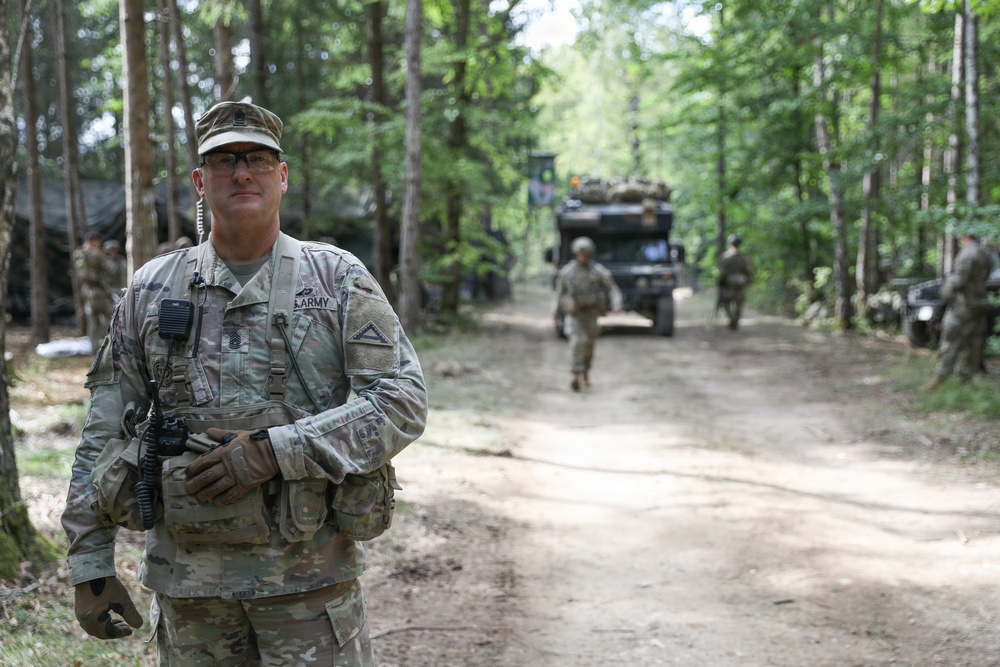 A U.S. Army soldier in MultiCam/OCP camouflage uniform stands in the foreground on a dirt forest road, holding a radio and wearing tactical gear. Behind him, additional soldiers and a large military truck are visible along the wooded trail. The scene appears to depict a field training exercise or operational deployment in a forested environment.