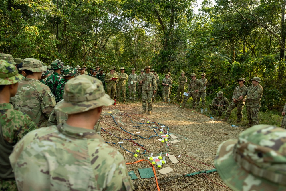A group of military personnel from multiple nations are gathered in an outdoor jungle clearing for what appears to be a tactical briefing or training exercise. A central figure is addressing the assembled soldiers around a ground-level terrain model or sand table constructed with ropes, markers, and small objects. The mixed uniforms suggest this is a multinational military exercise or joint training event.