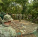 A group of military personnel from multiple nations are gathered in an outdoor jungle clearing for what appears to be a tactical briefing or training exercise. A central figure is addressing the assembled soldiers around a ground-level terrain model or sand table constructed with ropes, markers, and small objects. The mixed uniforms suggest this is a multinational military exercise or joint training event.