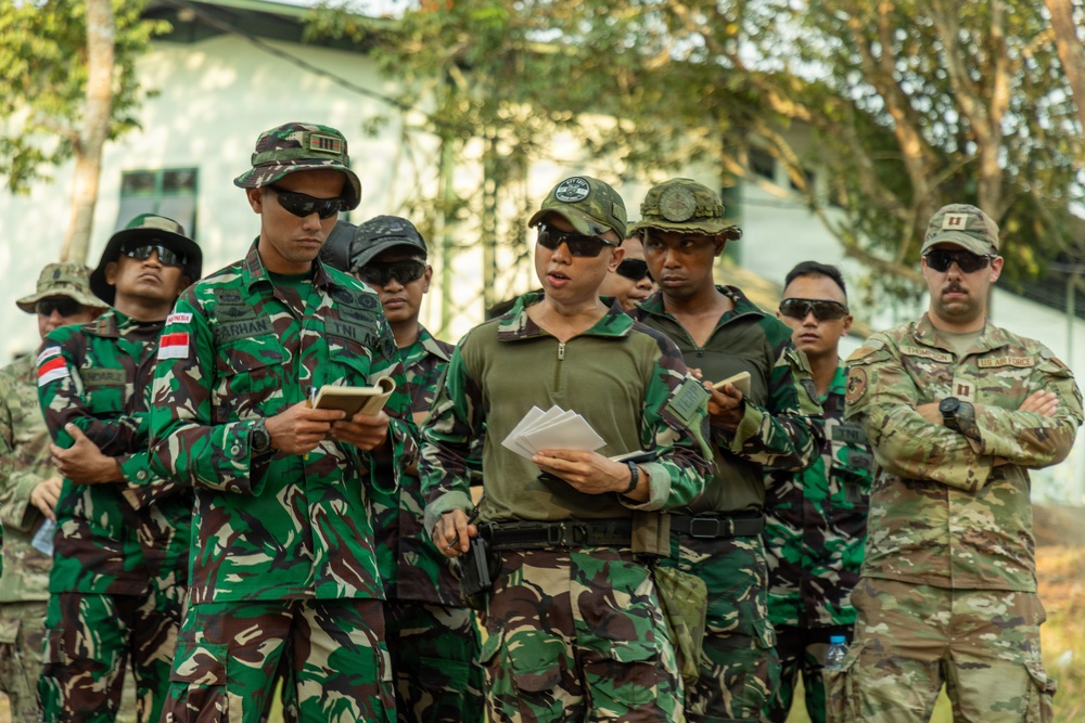 A group of military personnel from multiple nations are gathered outdoors, appearing to be engaged in a briefing or training exercise. Several soldiers are wearing Indonesian Army (TNI-AD) camouflage uniforms with visible name tapes and Indonesian flag patches, while at least one individual on the right wears a US Air Force multicam uniform with captain rank insignia. Personnel are reviewing documents and notes, suggesting a joint multinational military exercise or training event.