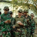 A group of military personnel from multiple nations are gathered outdoors, appearing to be engaged in a briefing or training exercise. Several soldiers are wearing Indonesian Army (TNI-AD) camouflage uniforms with visible name tapes and Indonesian flag patches, while at least one individual on the right wears a US Air Force multicam uniform with captain rank insignia. Personnel are reviewing documents and notes, suggesting a joint multinational military exercise or training event.