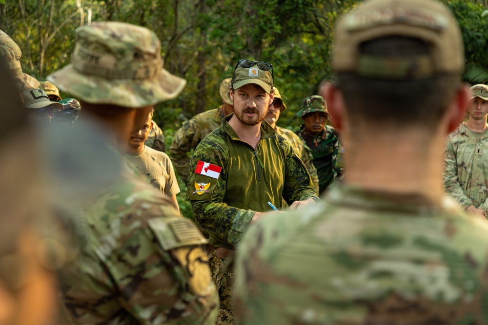 A multinational military gathering in an outdoor, wooded setting where a Canadian Armed Forces soldier is addressing a mixed group of military personnel. The central figure wears Canadian CADPAT camouflage with a visible Canadian flag patch and a Carhartt cap with sunglasses. The group includes personnel in various camouflage patterns suggesting participation from multiple nations.