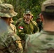 A multinational military gathering in an outdoor, wooded setting where a Canadian Armed Forces soldier is addressing a mixed group of military personnel. The central figure wears Canadian CADPAT camouflage with a visible Canadian flag patch and a Carhartt cap with sunglasses. The group includes personnel in various camouflage patterns suggesting participation from multiple nations.