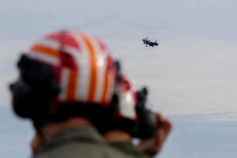 A military flight deck crew member wearing a distinctive red and orange striped helmet is photographing or observing a fixed-wing military jet aircraft in flight. The aircraft, visible in the background and in sharp focus, appears to be an F-35 variant based on its silhouette, with landing gear deployed suggesting an approach or departure. The foreground figure is intentionally blurred, drawing visual emphasis to the aircraft in the distance.