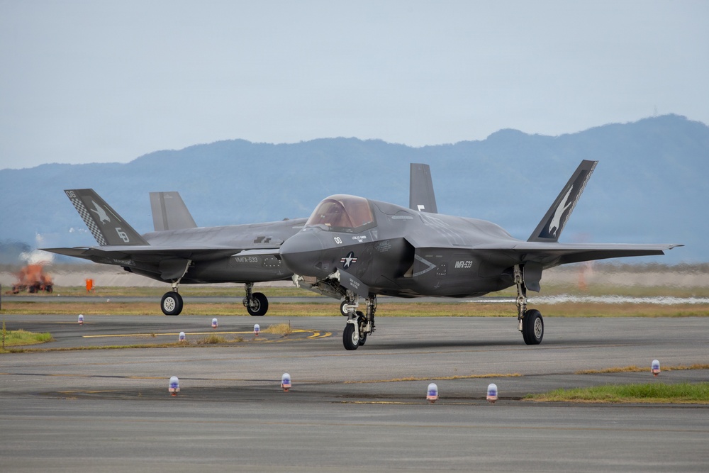Two F-35B stealth fighter jets assigned to VMFA-533 (Marine Fighter Attack Squadron 533) are taxiing on a runway apron. The aircraft display distinctive tail markings including a checkered pattern and a bird/hawk insignia. The setting appears to be a military airfield with mountains visible in the background.