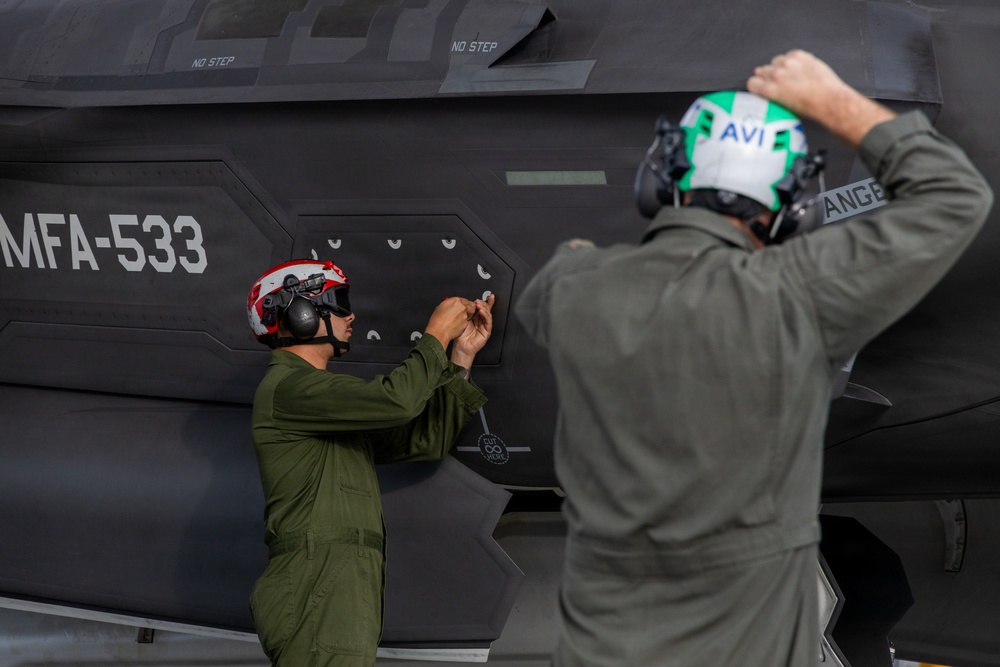 Two military personnel in olive drab flight suits are working on the fuselage of a military jet aircraft marked 'MFA-533'. One technician wearing a red helmet is manipulating fasteners or panels on the aircraft's side, while a second person in a green and white helmet labeled 'AVI' assists. The aircraft displays 'NO STEP' markings and has a dark stealth-style finish consistent with an F-35 series aircraft.