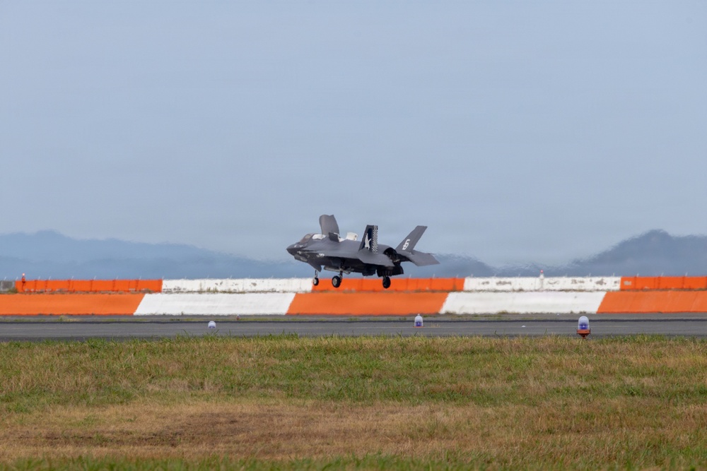 A military stealth fighter jet, identifiable as an F-35, is captured in a low-altitude approach or landing phase on a runway. The aircraft is photographed from a ground-level perspective with landing gear deployed. The background features a distinctive orange and white barrier wall, green grass infield, and mountainous terrain under a hazy sky.