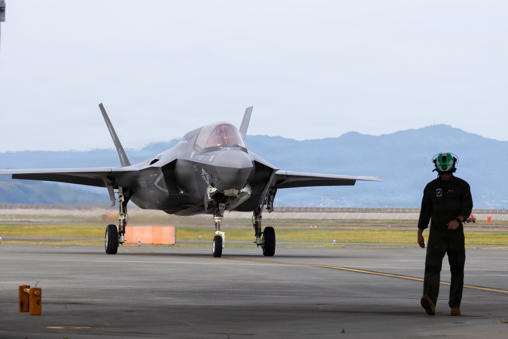 A fifth-generation stealth fighter jet, identifiable as an F-35, is taxiing on a military airfield runway. A ground crew member wearing a green helmet and dark flight suit is walking alongside the aircraft, likely performing marshalling or inspection duties. Mountains and a hazy sky are visible in the background.