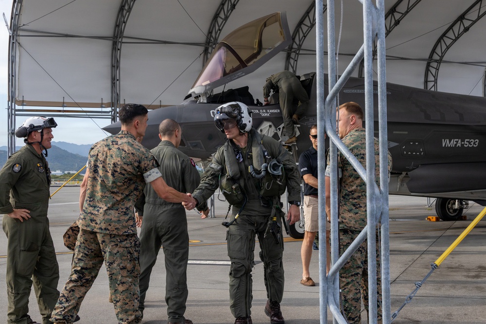 A military scene on an airfield tarmac showing personnel from what appears to be the U.S. Marine Corps gathered around an F-35 fighter jet. A pilot in full flight gear, including a helmet, is shaking hands with personnel in camouflage uniforms, suggesting a post-flight greeting or debrief. The aircraft is marked with 'VMFA-533' and a pilot name 'LTCOL D.R. KANSIER' with callsign 'DORA'.