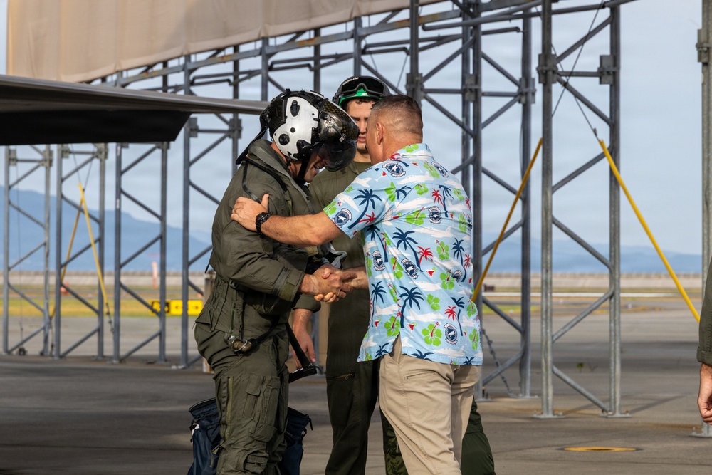 A military pilot in full flight gear, including a helmet with visor, is being greeted with a handshake by a civilian wearing a tropical Hawaiian-style shirt on an airfield tarmac. A second person in olive drab flight suit stands in the background observing the exchange. The setting appears to be a military air base, with large hangar framework structures visible behind the group.