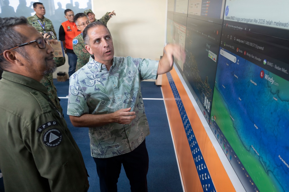 A group of officials are gathered in what appears to be an emergency management or disaster response operations center in Indonesia. A man in a floral shirt is pointing at a large display screen showing the Windy.com weather/meteorological interface, while a man wearing a BPBD (Badan Penanggulangan Bencana Daerah - Indonesian Disaster Management Agency) uniform listens attentively. Several other officials in various uniforms are visible in the background.