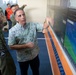 A group of officials are gathered in what appears to be an emergency management or disaster response operations center in Indonesia. A man in a floral shirt is pointing at a large display screen showing the Windy.com weather/meteorological interface, while a man wearing a BPBD (Badan Penanggulangan Bencana Daerah - Indonesian Disaster Management Agency) uniform listens attentively. Several other officials in various uniforms are visible in the background.