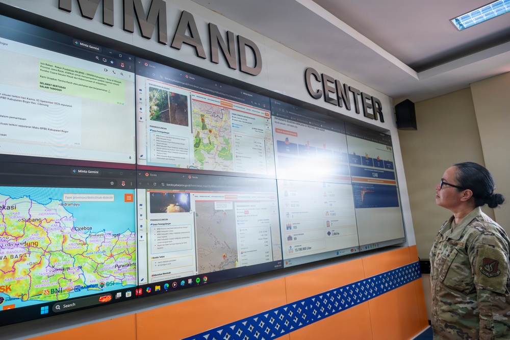 A person in military camouflage uniform stands before a large multi-screen video wall in a Command Center, appearing to review displayed information. The screens show various data dashboards, maps, messaging applications, and what appears to be disaster management or emergency response information in Indonesian language. The facility appears to be an Indonesian emergency management command center (BPBD - Badan Penanggulangan Bencana Daerah).