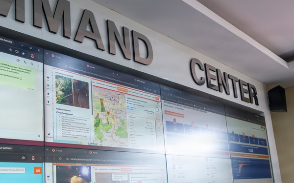 A person in military camouflage uniform stands before a large multi-screen video wall in a Command Center, appearing to review displayed information. The screens show various data dashboards, maps, messaging applications, and what appears to be disaster management or emergency response information in Indonesian language. The facility appears to be an Indonesian emergency management command center (BPBD - Badan Penanggulangan Bencana Daerah).