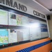 A person in military camouflage uniform stands before a large multi-screen video wall in a Command Center, appearing to review displayed information. The screens show various data dashboards, maps, messaging applications, and what appears to be disaster management or emergency response information in Indonesian language. The facility appears to be an Indonesian emergency management command center (BPBD - Badan Penanggulangan Bencana Daerah).