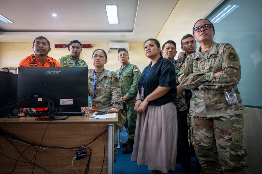 A group of military and civilian personnel from multiple nations are gathered in what appears to be an operations or command center, observing a computer monitor. The group includes individuals in different military uniforms including camouflage (OCP/Multicam pattern), a bright orange rescue uniform, and what appears to be Singapore Armed Forces attire, alongside civilians wearing lanyards. The setting suggests a multinational exercise, briefing, or coordination meeting.
