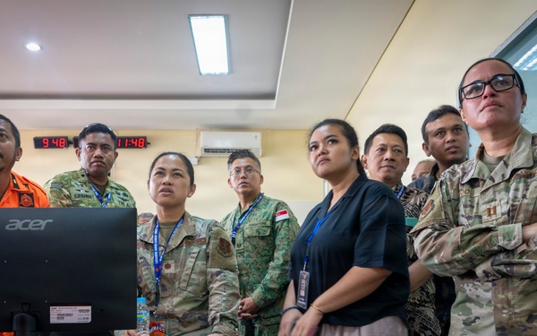 A group of military and civilian personnel from multiple nations are gathered in what appears to be an operations or command center, observing a computer monitor. The group includes individuals in different military uniforms including camouflage (OCP/Multicam pattern), a bright orange rescue uniform, and what appears to be Singapore Armed Forces attire, alongside civilians wearing lanyards. The setting suggests a multinational exercise, briefing, or coordination meeting.