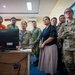 A group of military and civilian personnel from multiple nations are gathered in what appears to be an operations or command center, observing a computer monitor. The group includes individuals in different military uniforms including camouflage (OCP/Multicam pattern), a bright orange rescue uniform, and what appears to be Singapore Armed Forces attire, alongside civilians wearing lanyards. The setting suggests a multinational exercise, briefing, or coordination meeting.