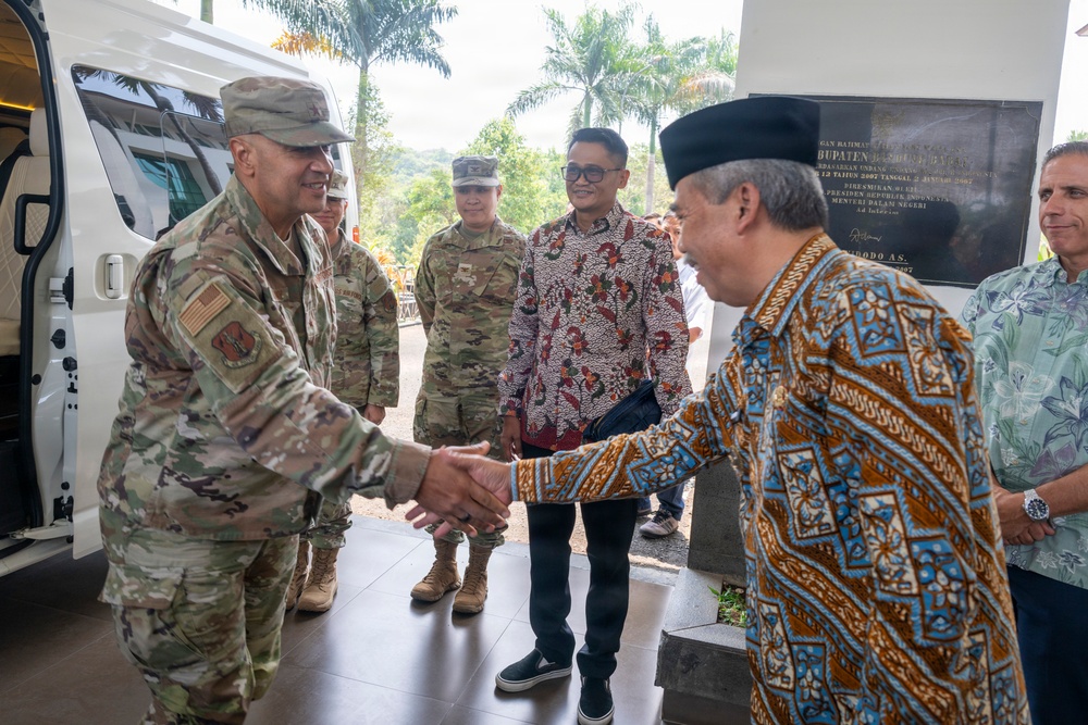 A formal greeting is taking place between U.S. military personnel in multicam/OCP camouflage uniforms and local officials wearing traditional Indonesian batik clothing. The setting appears to be an official visit or diplomatic engagement, likely in Indonesia, as evidenced by the batik attire and a commemorative plaque visible in the background. The handshake between a senior U.S. military officer and a local dignitary wearing a traditional black peci cap suggests a bilateral or diplomatic meeting.
