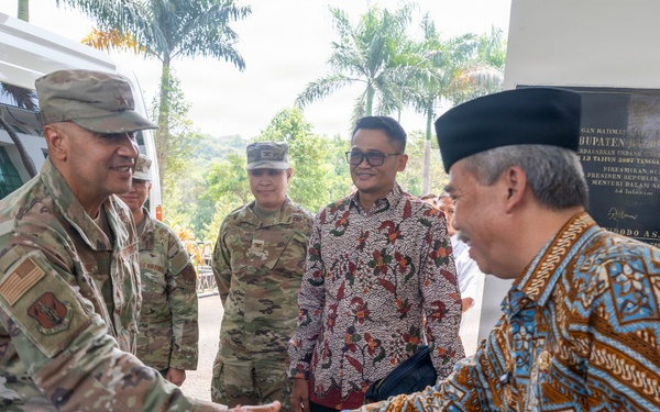 A formal greeting is taking place between U.S. military personnel in multicam/OCP camouflage uniforms and local officials wearing traditional Indonesian batik clothing. The setting appears to be an official visit or diplomatic engagement, likely in Indonesia, as evidenced by the batik attire and a commemorative plaque visible in the background. The handshake between a senior U.S. military officer and a local dignitary wearing a traditional black peci cap suggests a bilateral or diplomatic meeting.