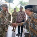 A formal greeting is taking place between U.S. military personnel in multicam/OCP camouflage uniforms and local officials wearing traditional Indonesian batik clothing. The setting appears to be an official visit or diplomatic engagement, likely in Indonesia, as evidenced by the batik attire and a commemorative plaque visible in the background. The handshake between a senior U.S. military officer and a local dignitary wearing a traditional black peci cap suggests a bilateral or diplomatic meeting.