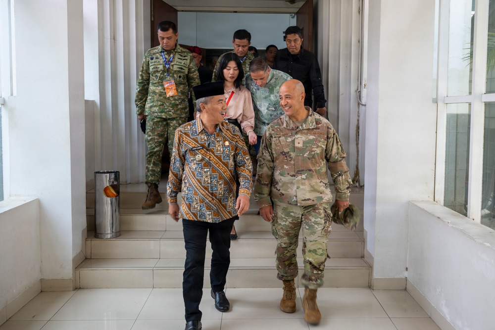 The image shows a group of individuals descending steps from a building, with two prominent figures in the foreground engaged in conversation. One foreground figure is wearing traditional Indonesian batik attire with a peci (black cap), while the other is in US Army MultiCam (OCP) combat uniform. Several other individuals in the background include military personnel in camouflage uniforms and civilians.