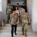The image shows a group of individuals descending steps from a building, with two prominent figures in the foreground engaged in conversation. One foreground figure is wearing traditional Indonesian batik attire with a peci (black cap), while the other is in US Army MultiCam (OCP) combat uniform. Several other individuals in the background include military personnel in camouflage uniforms and civilians.