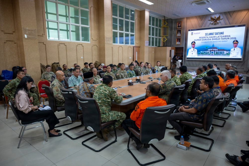 A multinational military briefing or coordination meeting is taking place in a conference room in Kabupaten Bandung Barat, Indonesia. Participants include military personnel in various uniforms (Indonesian, US, and other allied forces), civilians in traditional Indonesian attire, and individuals in orange vests likely representing disaster response agencies. A welcome screen displays 'Selamat Datang Peserta HADR LATGABMA Super Garuda Shield Tahun 2026,' indicating this is a Humanitarian Assistance and Disaster Relief (HADR) joint exercise briefing.