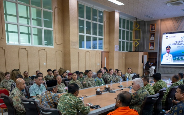 A multinational military briefing or coordination meeting is taking place in a conference room in Kabupaten Bandung Barat, Indonesia. Participants include military personnel in various uniforms (Indonesian, US, and other allied forces), civilians in traditional Indonesian attire, and individuals in orange vests likely representing disaster response agencies. A welcome screen displays 'Selamat Datang Peserta HADR LATGABMA Super Garuda Shield Tahun 2026,' indicating this is a Humanitarian Assistance and Disaster Relief (HADR) joint exercise briefing.
