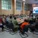 A multinational military briefing or coordination meeting is taking place in a conference room in Kabupaten Bandung Barat, Indonesia. Participants include military personnel in various uniforms (Indonesian, US, and other allied forces), civilians in traditional Indonesian attire, and individuals in orange vests likely representing disaster response agencies. A welcome screen displays 'Selamat Datang Peserta HADR LATGABMA Super Garuda Shield Tahun 2026,' indicating this is a Humanitarian Assistance and Disaster Relief (HADR) joint exercise briefing.