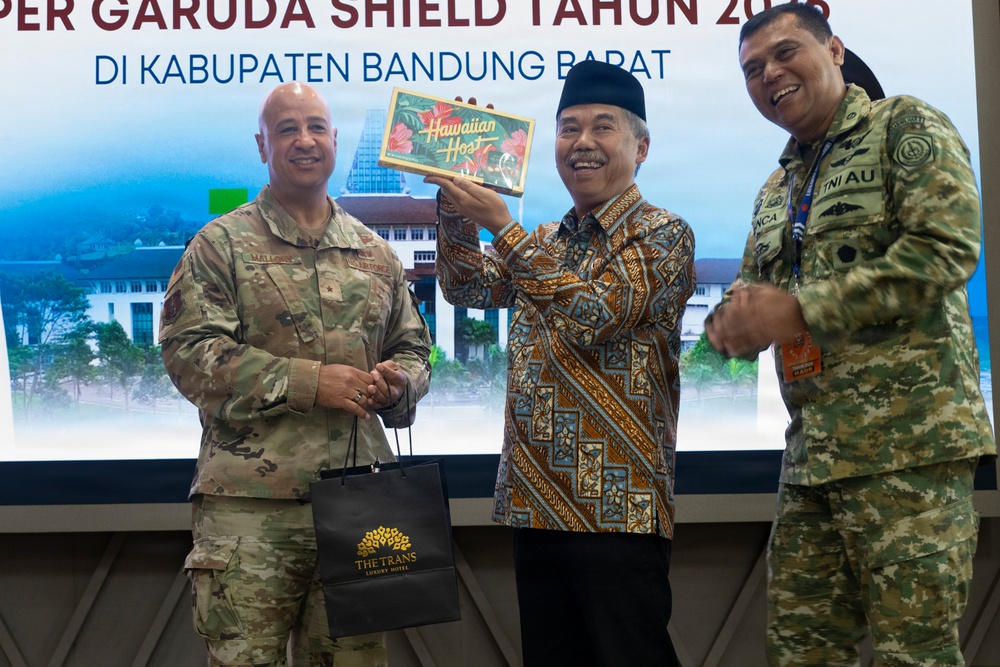 Three individuals are posing for a photo at an event related to 'Super Garuda Shield' in Kabupaten Bandung Barat, Indonesia. A civilian in traditional Indonesian batik clothing and a peci (black cap) is holding up a 'Hawaiian Host' gift box, flanked by two military personnel — one in US Army camouflage uniform and one in Indonesian TNI AU (Air Force) camouflage uniform. A 'The Trans Luxury Hotel' branded gift bag is also visible, suggesting a formal gift exchange ceremony.