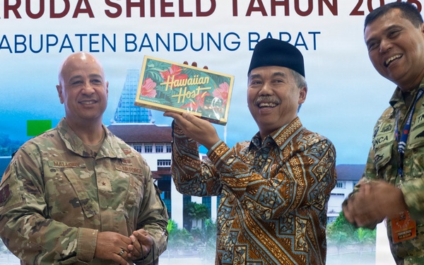 Three individuals are posing for a photo at an event related to 'Super Garuda Shield' in Kabupaten Bandung Barat, Indonesia. A civilian in traditional Indonesian batik clothing and a peci (black cap) is holding up a 'Hawaiian Host' gift box, flanked by two military personnel — one in US Army camouflage uniform and one in Indonesian TNI AU (Air Force) camouflage uniform. A 'The Trans Luxury Hotel' branded gift bag is also visible, suggesting a formal gift exchange ceremony.
