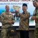 Three individuals are posing for a photo at an event related to 'Super Garuda Shield' in Kabupaten Bandung Barat, Indonesia. A civilian in traditional Indonesian batik clothing and a peci (black cap) is holding up a 'Hawaiian Host' gift box, flanked by two military personnel — one in US Army camouflage uniform and one in Indonesian TNI AU (Air Force) camouflage uniform. A 'The Trans Luxury Hotel' branded gift bag is also visible, suggesting a formal gift exchange ceremony.