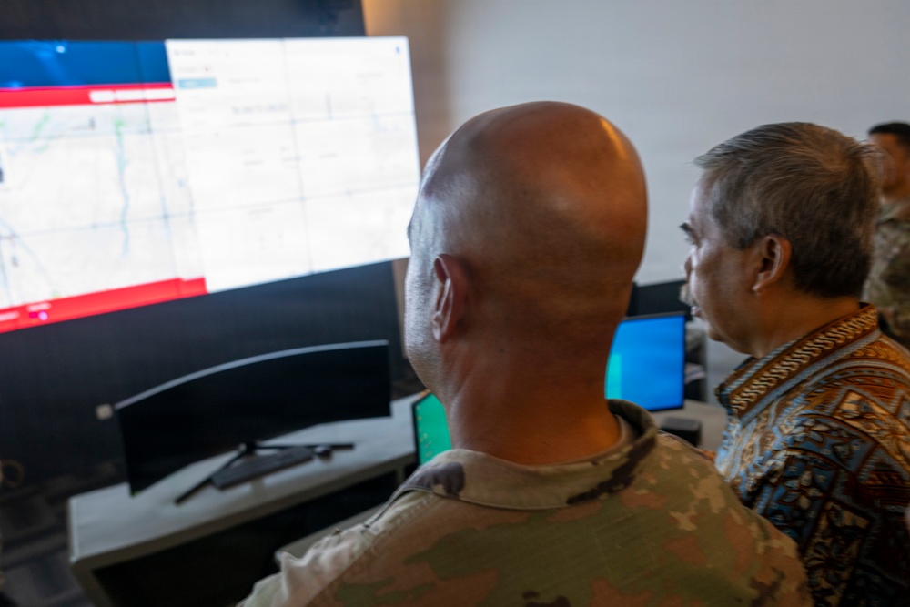 Two individuals are viewing a large curved monitor displaying what appears to be a digital map interface with a red banner element. One person is wearing military camouflage uniform and is bald, while the other is wearing a colorful patterned civilian shirt. The setting appears to be a command or operations center with multiple computer screens visible.