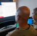 Two individuals are viewing a large curved monitor displaying what appears to be a digital map interface with a red banner element. One person is wearing military camouflage uniform and is bald, while the other is wearing a colorful patterned civilian shirt. The setting appears to be a command or operations center with multiple computer screens visible.