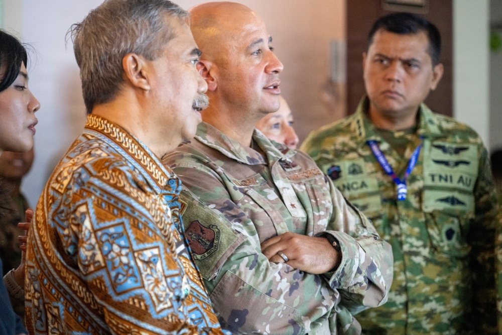 The image shows a group of military and civilian personnel gathered together, appearing to observe or listen to a presentation or briefing. At least two individuals are wearing military camouflage uniforms, one of which displays 'TNI AU' (Tentara Nasional Indonesia Angkatan Udara - Indonesian Air Force) insignia. A civilian in a traditional batik-patterned shirt stands alongside the uniformed personnel, suggesting a multinational or civil-military engagement.