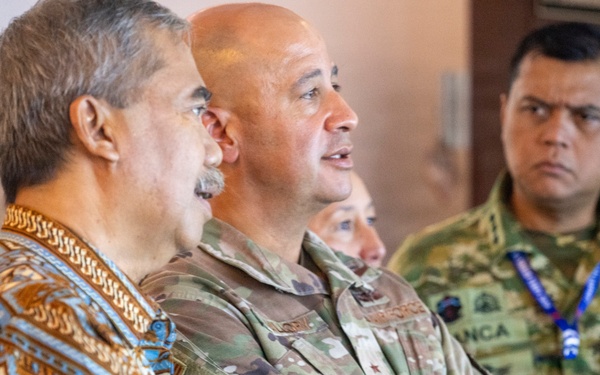 The image shows a group of military and civilian personnel gathered together, appearing to observe or listen to a presentation or briefing. At least two individuals are wearing military camouflage uniforms, one of which displays 'TNI AU' (Tentara Nasional Indonesia Angkatan Udara - Indonesian Air Force) insignia. A civilian in a traditional batik-patterned shirt stands alongside the uniformed personnel, suggesting a multinational or civil-military engagement.