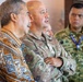 The image shows a group of military and civilian personnel gathered together, appearing to observe or listen to a presentation or briefing. At least two individuals are wearing military camouflage uniforms, one of which displays 'TNI AU' (Tentara Nasional Indonesia Angkatan Udara - Indonesian Air Force) insignia. A civilian in a traditional batik-patterned shirt stands alongside the uniformed personnel, suggesting a multinational or civil-military engagement.