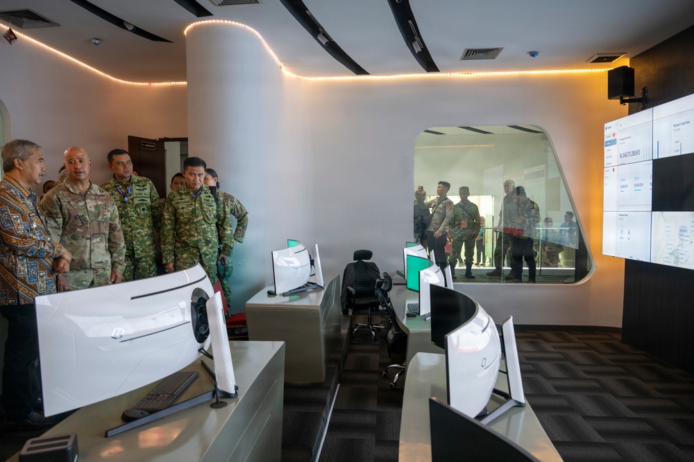 A group of military and civilian officials are touring a modern operations or command center facility. The room features multiple computer workstations with curved monitors, a large video wall display, and a distinctive oval observation window. Personnel in various military uniforms and civilian attire are observing the facility, with additional observers visible through the glass window.
