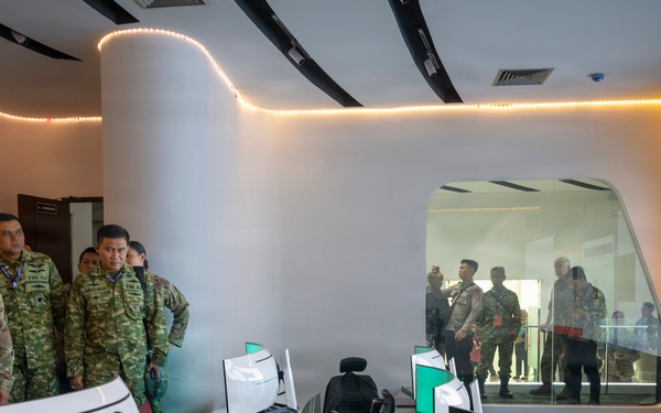 A group of military and civilian officials are touring a modern operations or command center facility. The room features multiple computer workstations with curved monitors, a large video wall display, and a distinctive oval observation window. Personnel in various military uniforms and civilian attire are observing the facility, with additional observers visible through the glass window.