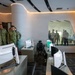 A group of military and civilian officials are touring a modern operations or command center facility. The room features multiple computer workstations with curved monitors, a large video wall display, and a distinctive oval observation window. Personnel in various military uniforms and civilian attire are observing the facility, with additional observers visible through the glass window.