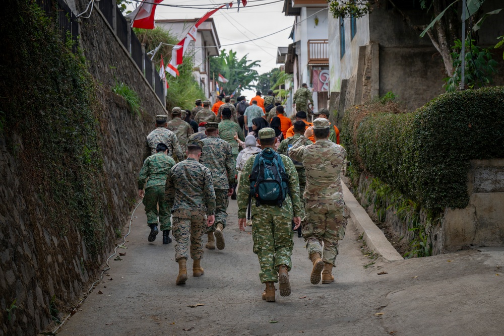 A group of military personnel in various camouflage uniforms, along with civilians in orange vests, are walking together down a narrow residential alleyway. Red and white flags are strung overhead, consistent with Indonesian national colors. The scene appears to depict a joint civil-military activity or community engagement operation in an urban village setting.