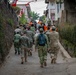 A group of military personnel in various camouflage uniforms, along with civilians in orange vests, are walking together down a narrow residential alleyway. Red and white flags are strung overhead, consistent with Indonesian national colors. The scene appears to depict a joint civil-military activity or community engagement operation in an urban village setting.